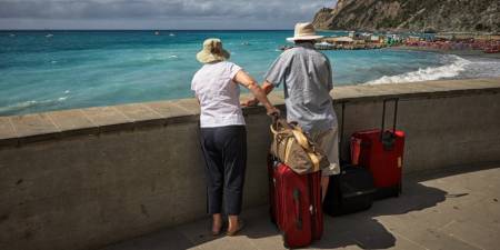 An elderly couple standing by the sea with suitcases.